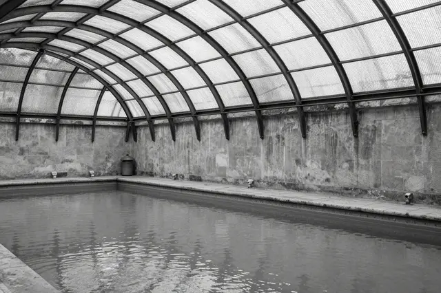 Black and white view of an empty indoor public bathhouse with an arched iron and glass roof over still pool water