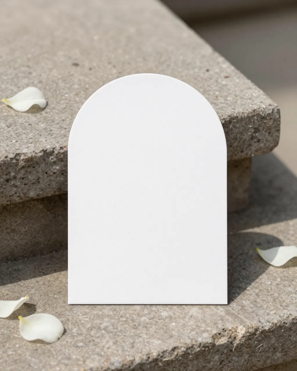 Blank arched white card sits on a stacked stone plinth with scattered white rose petals in natural sunlight