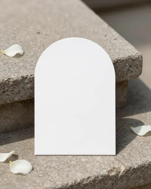 Blank arched white card sits on a stacked stone plinth with scattered white rose petals in natural sunlight