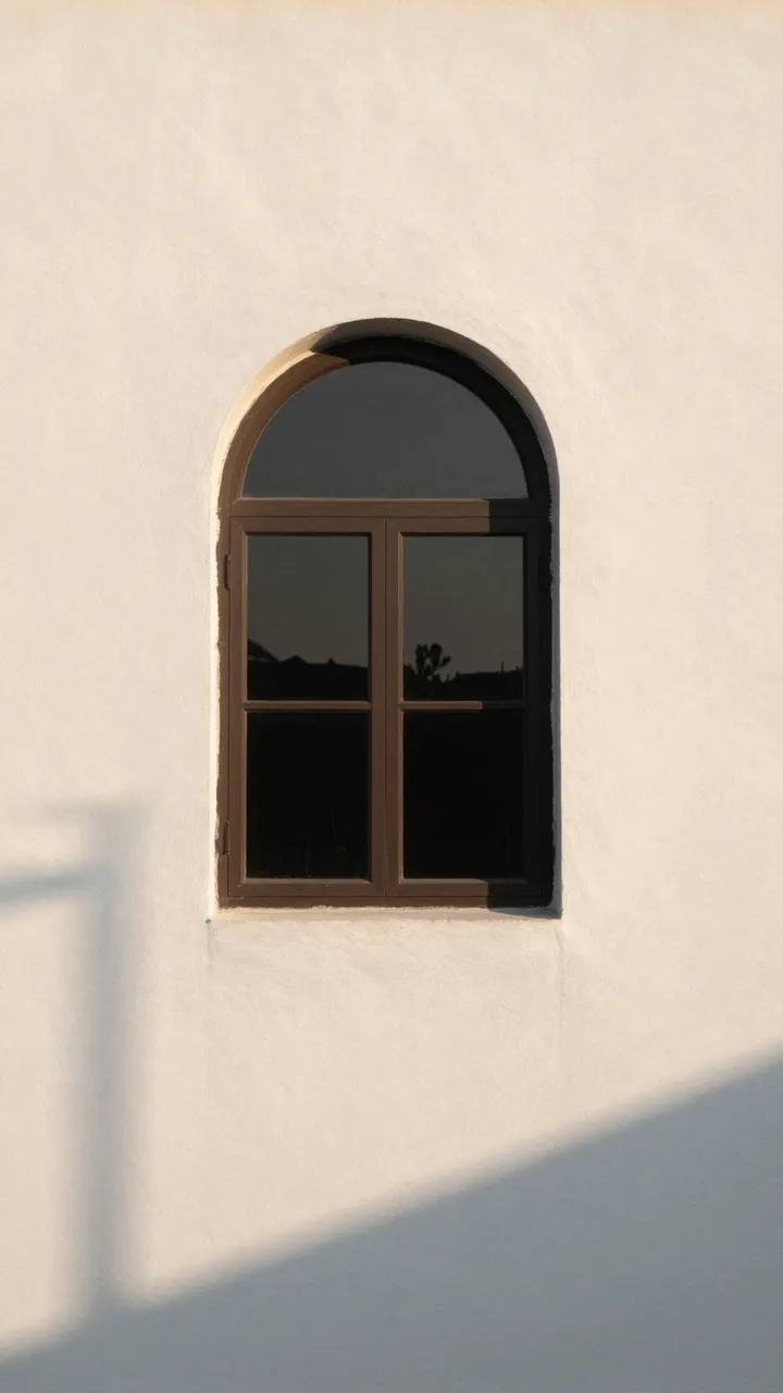 An arched dark framed window sits on a textured white plaster wall with soft golden hour shadows