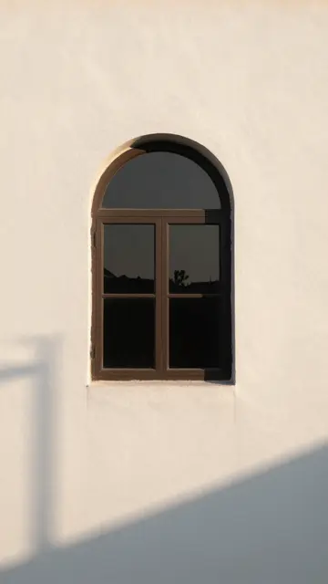 An arched dark framed window sits on a textured white plaster wall with soft golden hour shadows