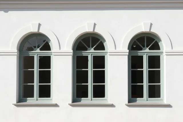 Three matching arched windows with pale green frames set in a smooth white stucco building facade