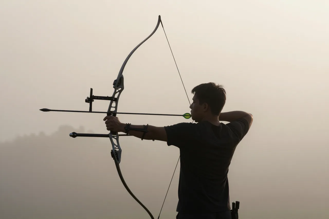 An adult archer draws a bow while aiming out into pale hazy morning fog