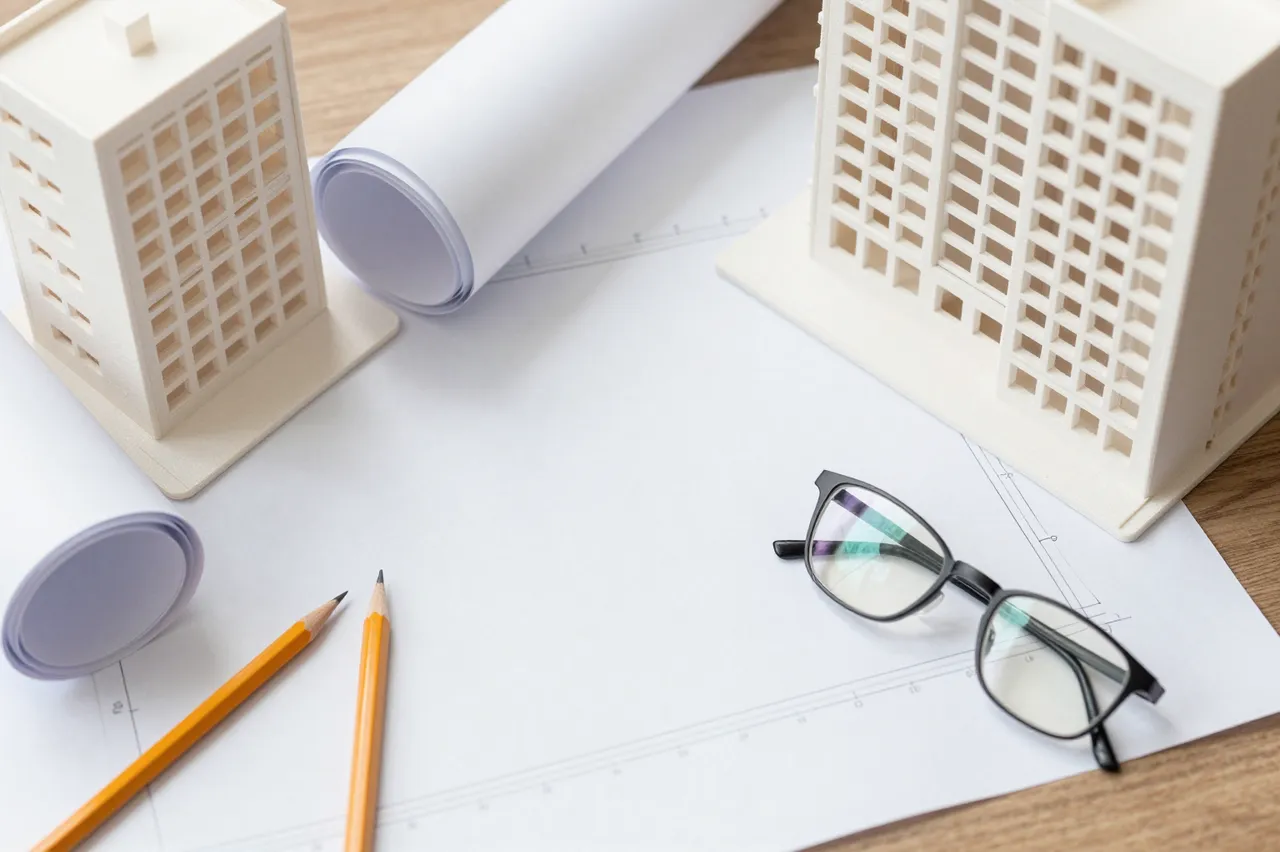 Top-down view of an architect's wooden desk holding building models, rolled drafting paper, pencils, and eyeglasses over a