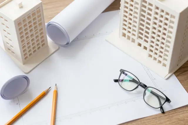 Top-down view of an architect's wooden desk holding building models, rolled drafting paper, pencils, and eyeglasses over a