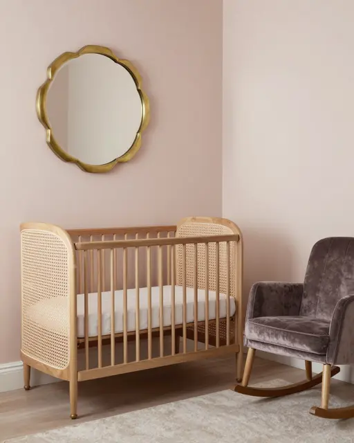 Empty art deco nursery with a cane crib, scalloped brass mirror, and velvet rocking chair against pale pink walls