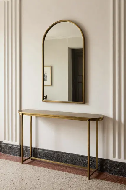 An arched brass mirror hangs above a curved brass console table against a fluted plaster wall with terrazzo flooring