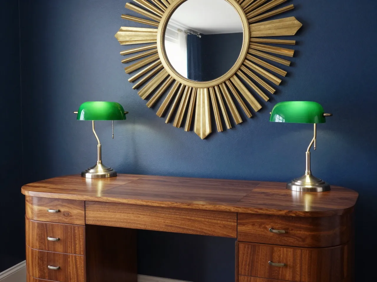 Curved walnut desk with green glass lamps sits below a gold sunburst mirror on navy walls