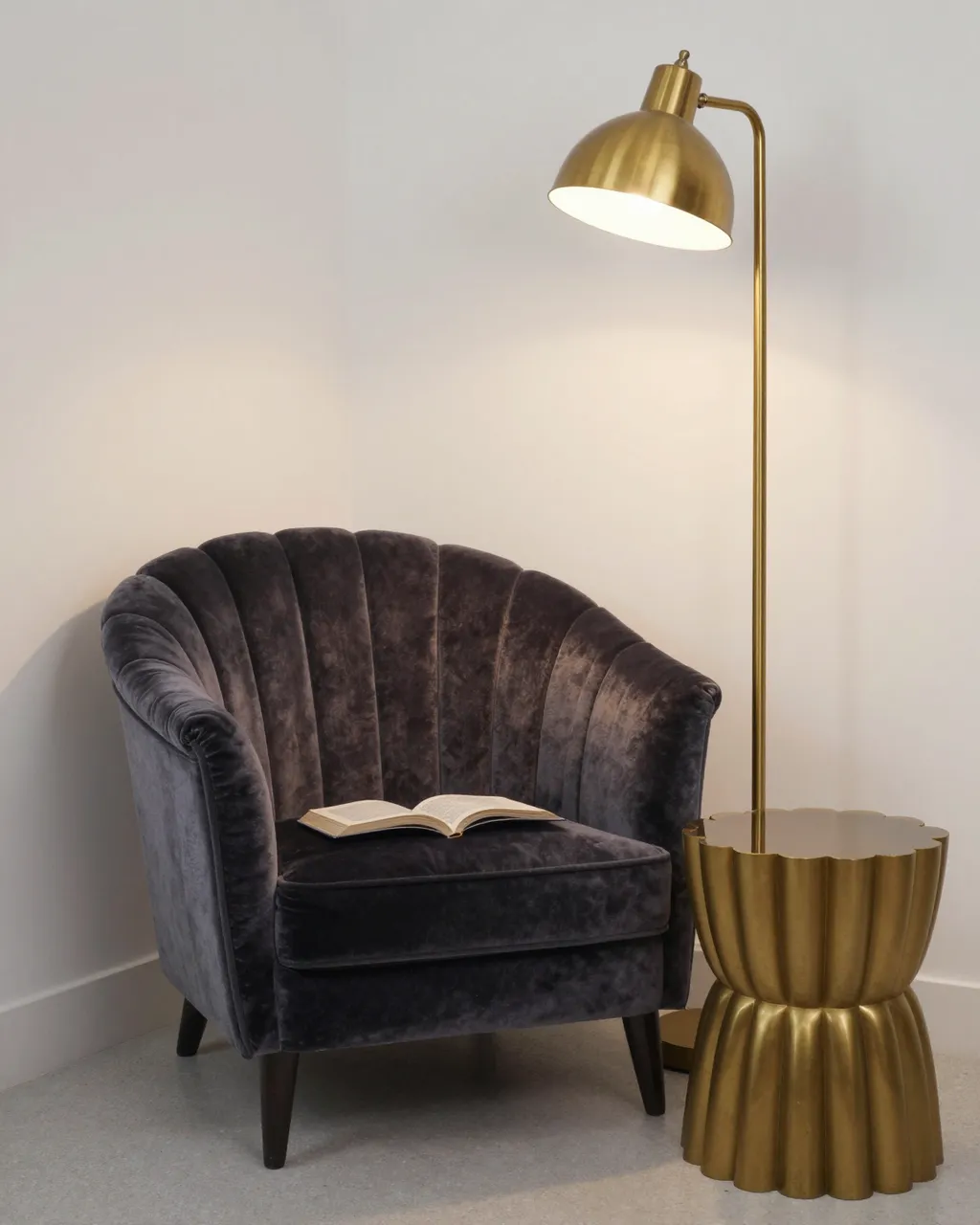 A dark velvet shell-back armchair with an open book sits beside a brass floor lamp and scalloped side table in a light