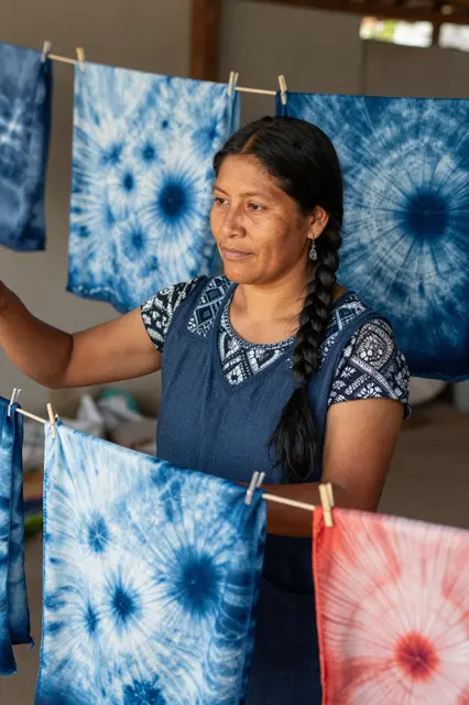 Indigenous female artisan stands among tie-dyed scarves drying on wooden lines in her studio
