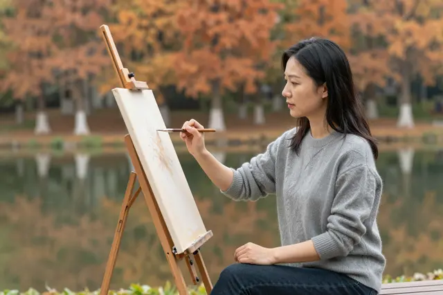East Asian woman in a gray sweater paints at an easel beside an autumn park pond