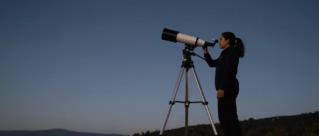 Woman looks through a large telescope set up on a hilltop under a fading dusk sky