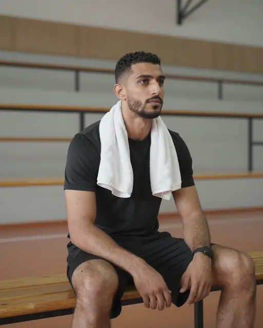 Middle Eastern man rests on a gym bench with a towel around his neck in an empty sports hall