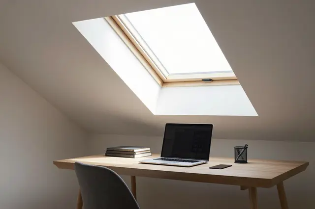 Sunlight from a sloped ceiling skylight illuminates a wooden home office desk with a laptop, books and chair.