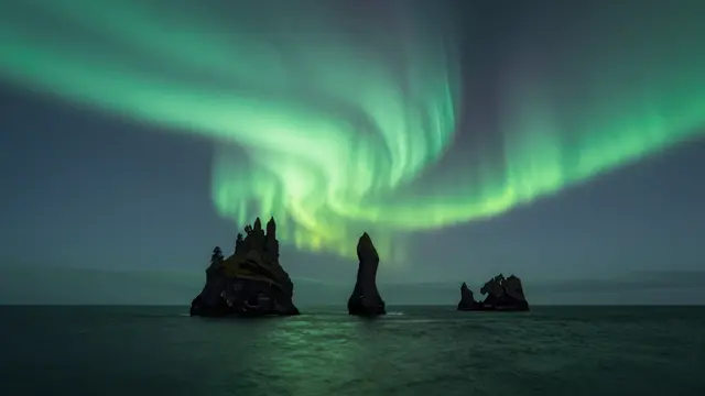 Swirling green northern lights glow over jagged sea stacks rising from a dark calm night ocean