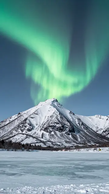Green aurora arc glows above a snowy mountain and frozen lake under a clear sky
