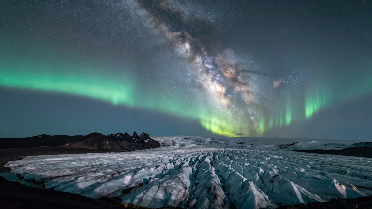 Bright green aurora and the glowing Milky Way stretch across the night sky above a crevassed glacier