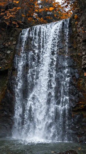 A cascading waterfall flows over dark rock framed by orange autumn tree foliage into a pool below