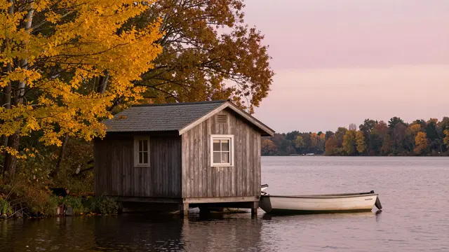 Weathered wooden boathouse sits on a calm lakeshore beside golden fall foliage at pink dusk