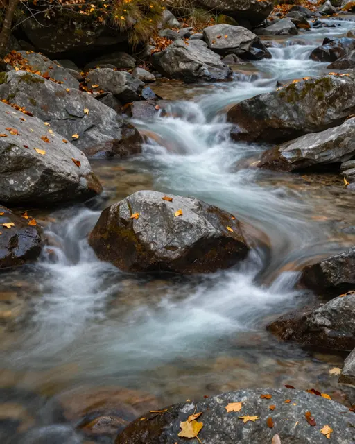 Silky flowing mountain stream parts around a large rounded boulder scattered with fallen autumn leaves