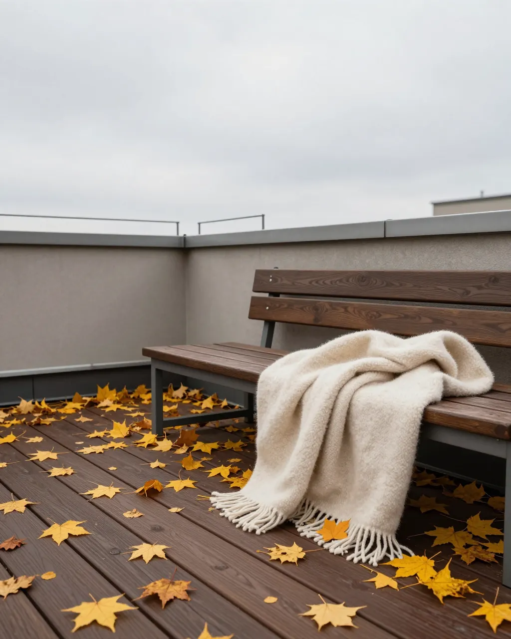 A wooden rooftop bench draped with a cream wool blanket sits scattered with fallen yellow maple leaves under an overcast sky
