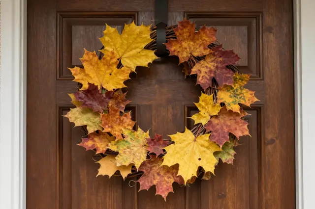 A fall maple leaf wreath hangs from a black strap on a dark brown paneled wooden front door.