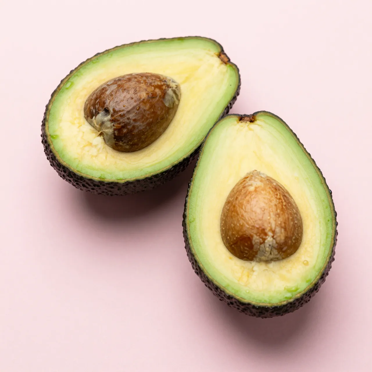 Top-down view of two halved avocados with pits resting on a soft pastel pink surface
