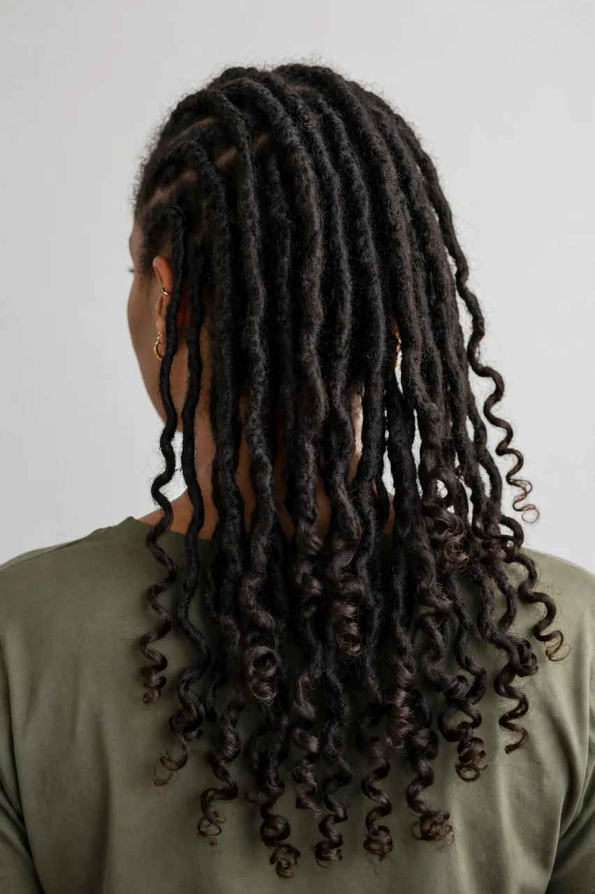 Back view of a woman showing long goddess locs with curly ends against a plain light backdrop