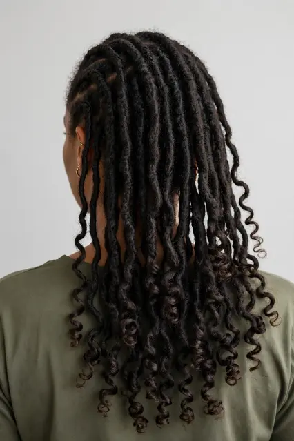 Back view of a woman showing long goddess locs with curly ends against a plain light backdrop