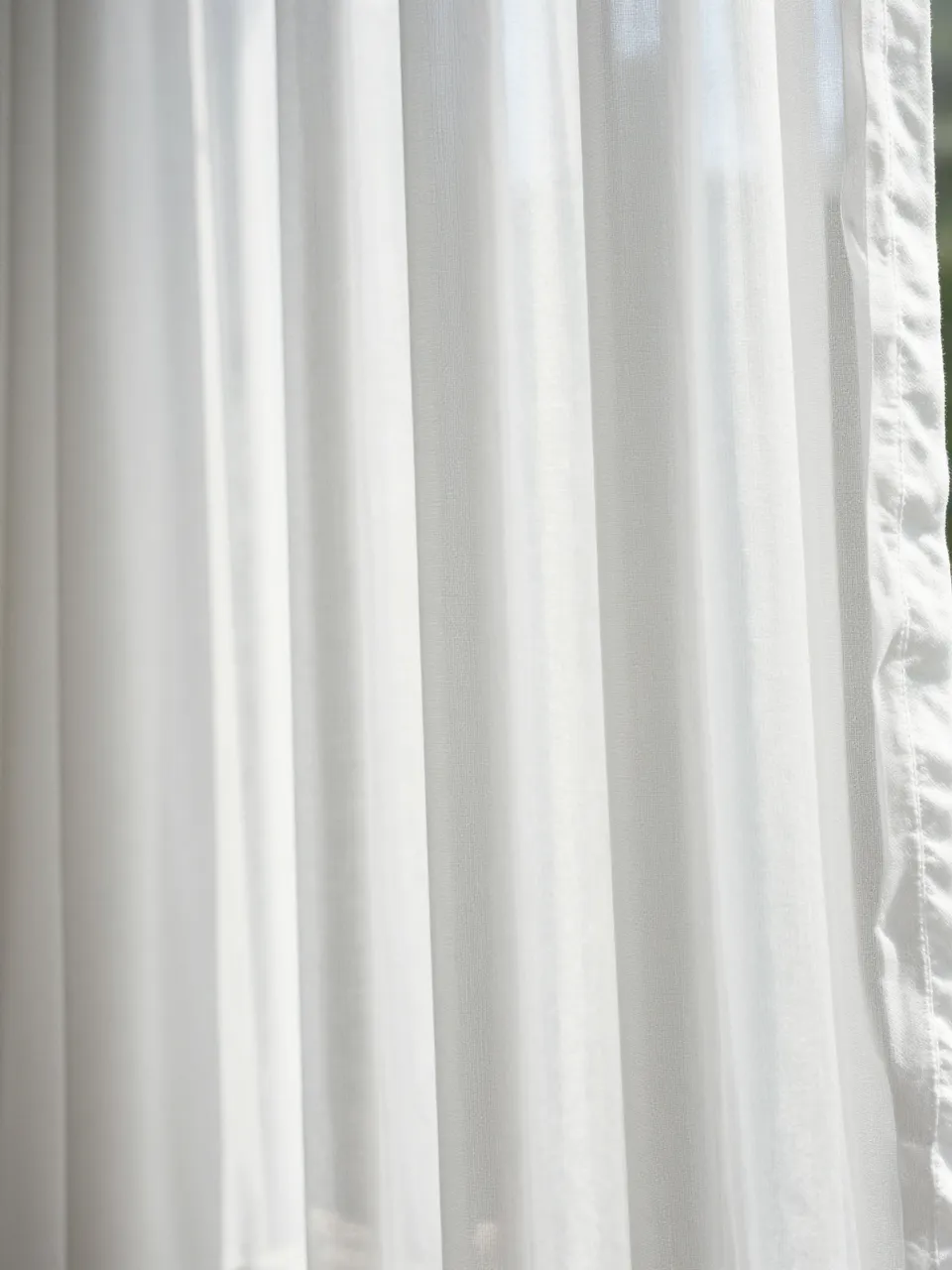 Full frame close-up of backlit white cotton voile sheer curtain with soft vertical folds.