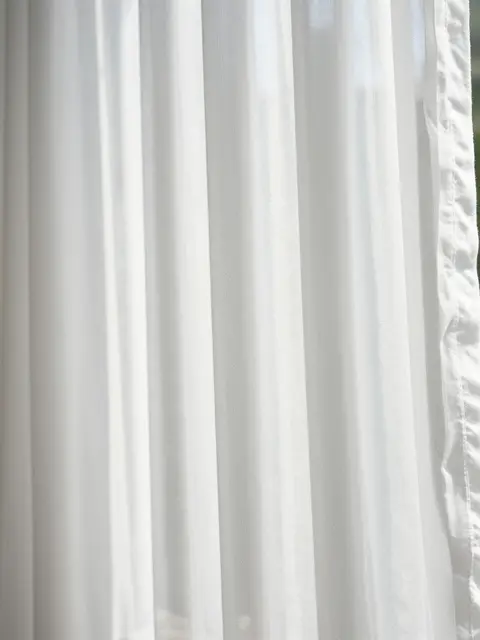 Full frame close-up of backlit white cotton voile sheer curtain with soft vertical folds.