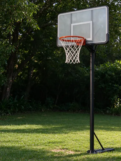 A portable basketball hoop stands on a sun-dappled backyard lawn shaded by trees at morning