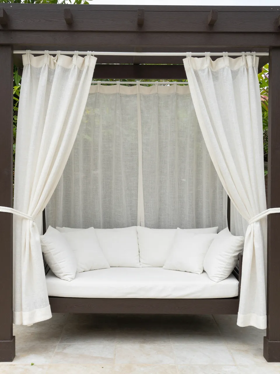 Dark wood backyard pergola holding a hanging daybed with white cushions and sheer linen curtains