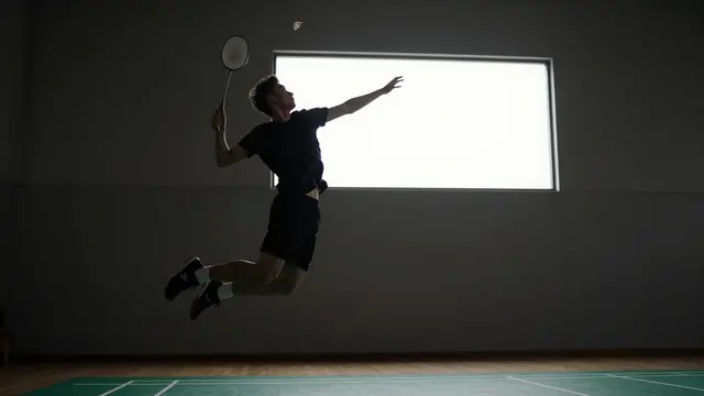 Silhouetted adult badminton player mid-jump to hit a shuttlecock in a dim indoor hall with a bright rear window