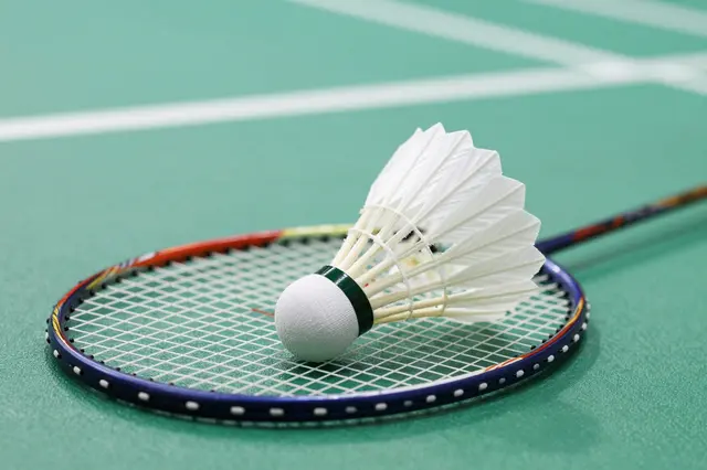 A badminton shuttlecock rests centered on the strings of a racket placed on a green indoor court.
