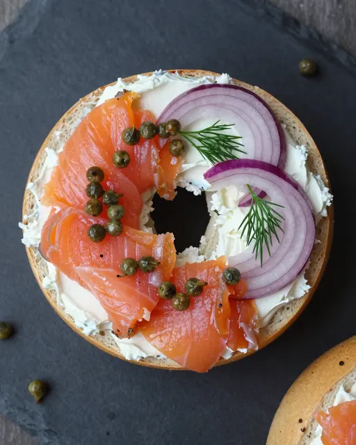 Top-down flat lay of a bagel topped with cream cheese, smoked salmon, red onion, capers, and dill on a dark slate board.
