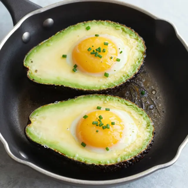 Two baked avocado halves with cooked egg and chive sprinkles sit in a small black skillet