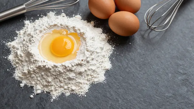 Top view of flour with a cracked egg, whole brown eggs, and whisks arranged on a dark slate table with empty copy space on