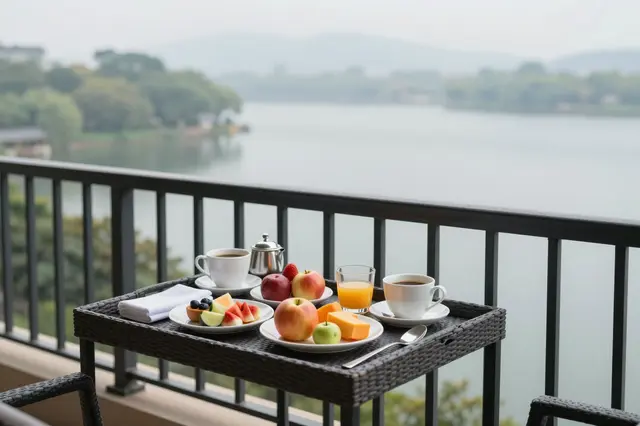 A breakfast tray with fruit, coffee and orange juice sits on a hotel balcony overlooking a hazy calm lake at morning