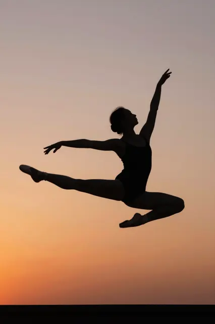 Dark silhouette of a leaping ballet dancer against a soft orange and pink gradient sunset sky