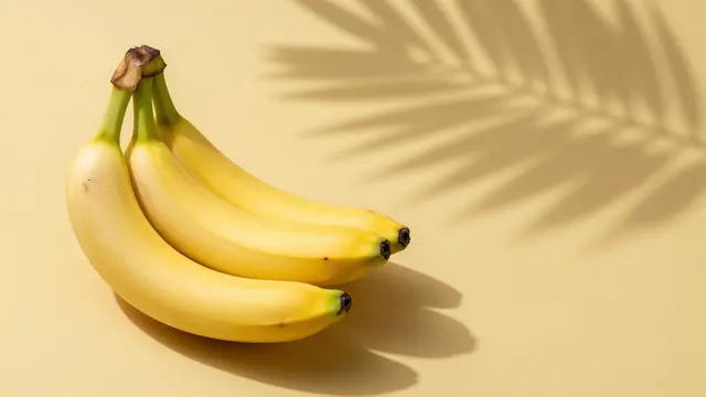 A bunch of ripe yellow bananas rests on a pale yellow surface with crisp palm leaf shadow and empty copy space to the right.