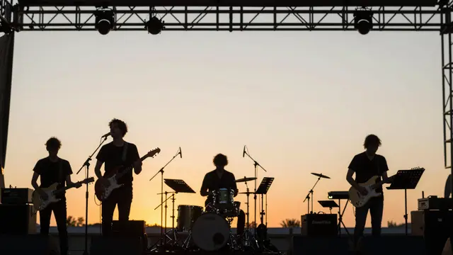 Silhouetted band members perform on an outdoor stage against a warm sunset sky