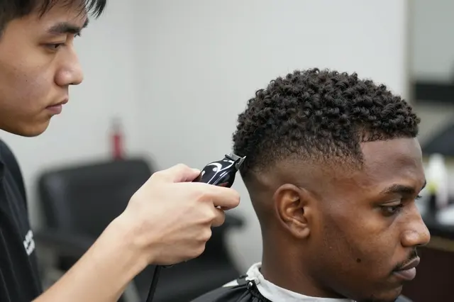 An East Asian barber uses clippers to fade the side of a Black client's haircut in a barbershop side view.