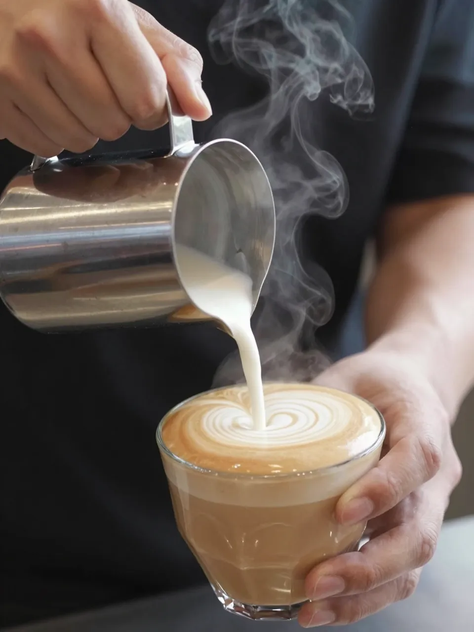 A barista pours steamed milk from a metal pitcher into a glass latte cup to create latte art, with rising steam.