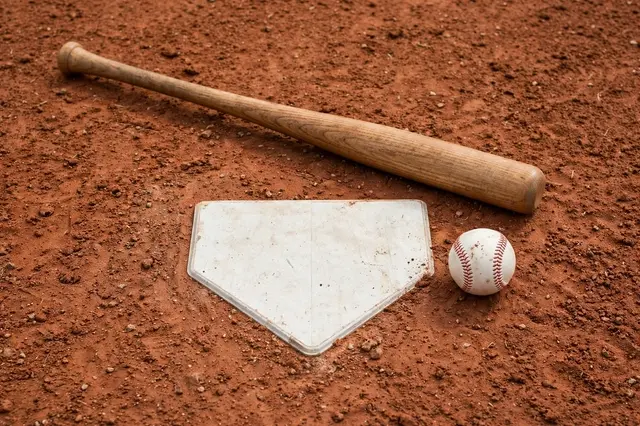 Wooden baseball bat, white home plate, and stitched baseball rest on red infield dirt