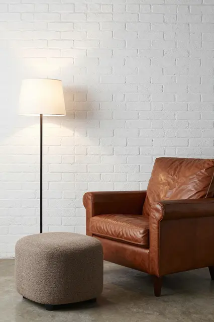Brown leather armchair, upholstered ottoman and floor lamp sit against a whitewashed brick wall