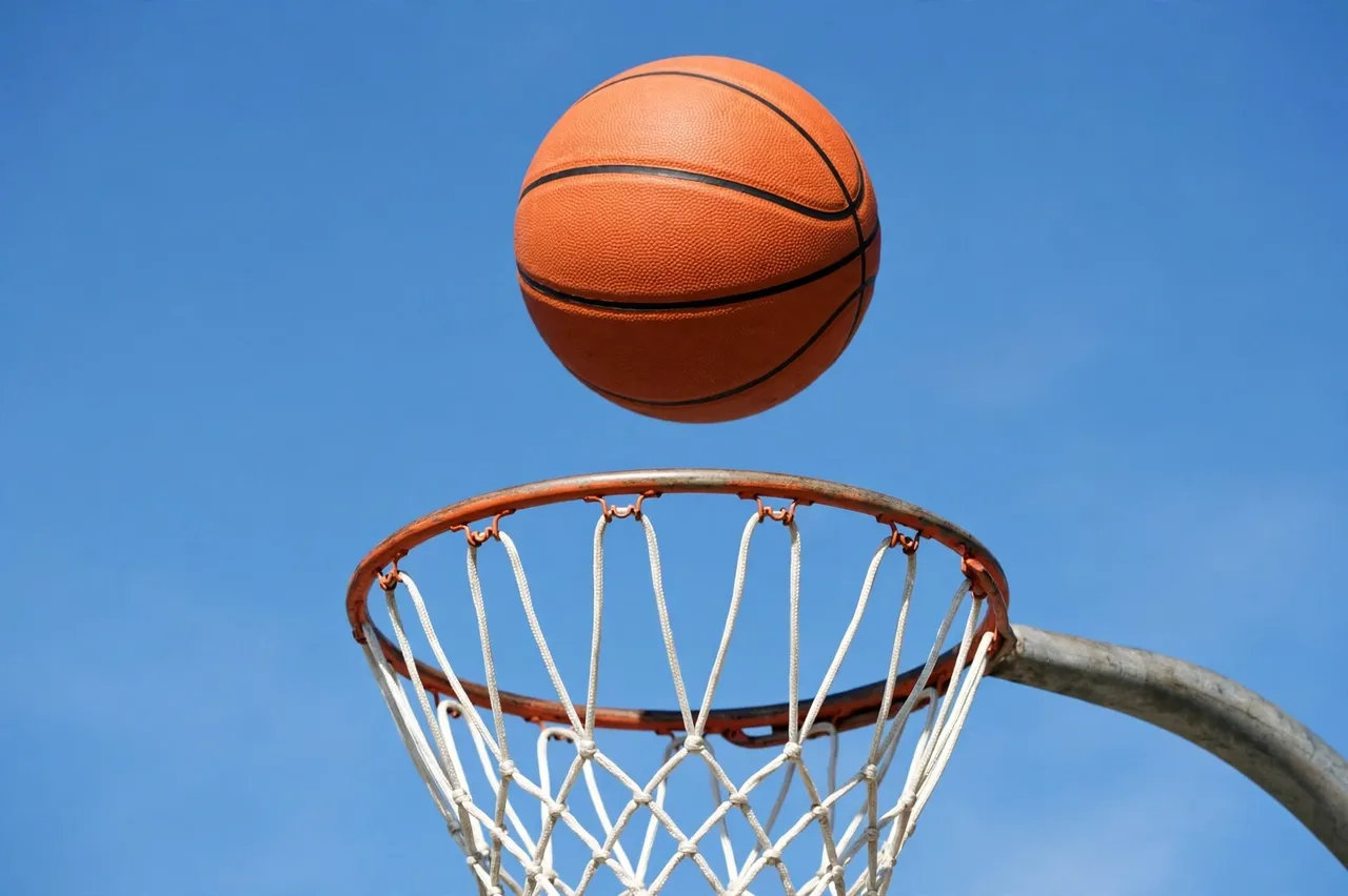 An orange basketball floats mid-air just above a basketball hoop against a clear bright blue sky.