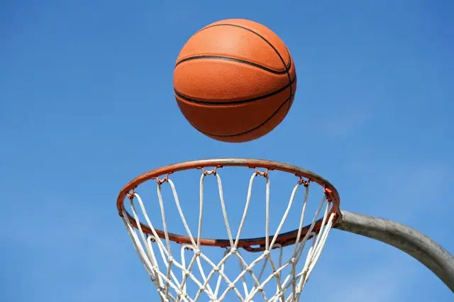 An orange basketball floats mid-air just above a basketball hoop against a clear bright blue sky.