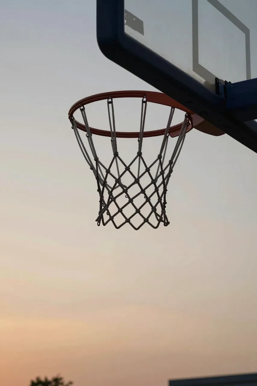 A basketball hoop with chain net and plain backboard is silhouetted against a soft gradient sunset sky.