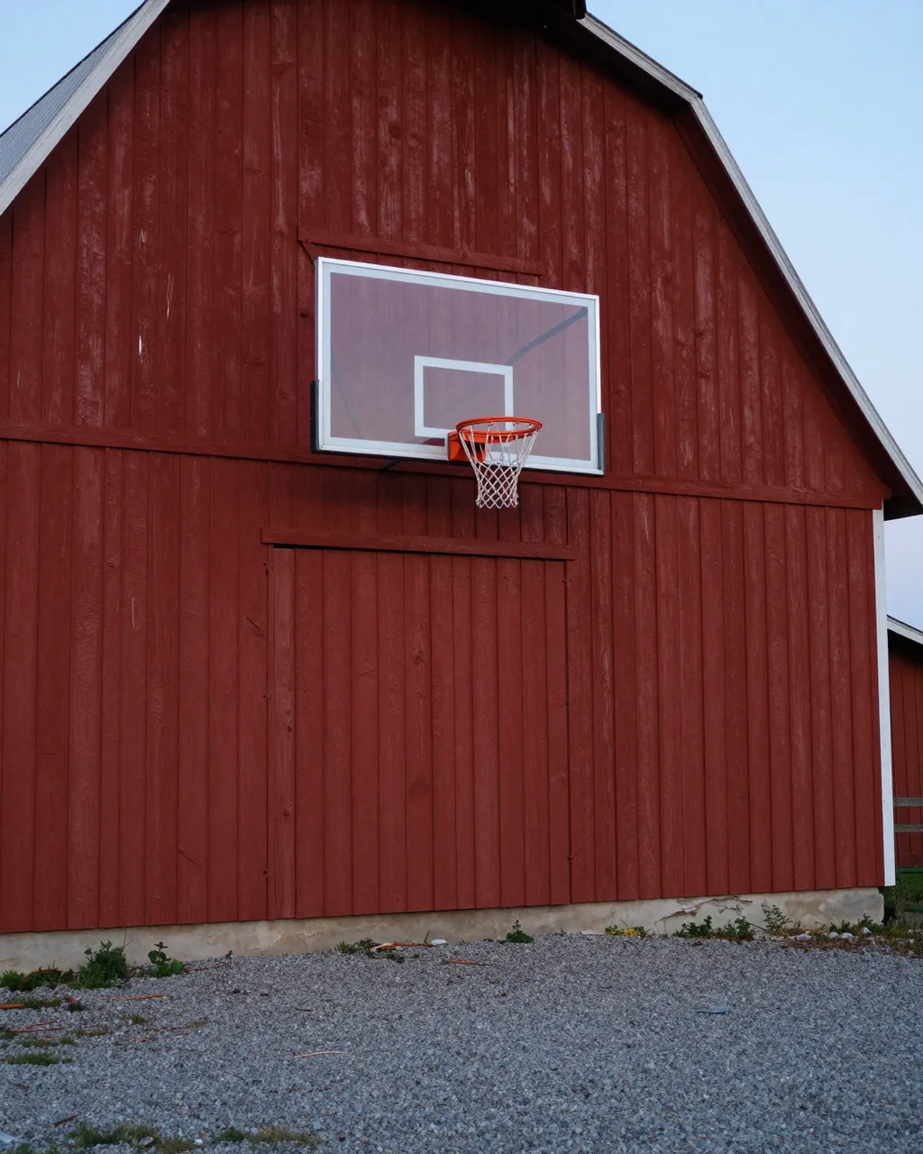 A basketball hoop mounted on the wall of an old red barn above a gravel yard at dusk
